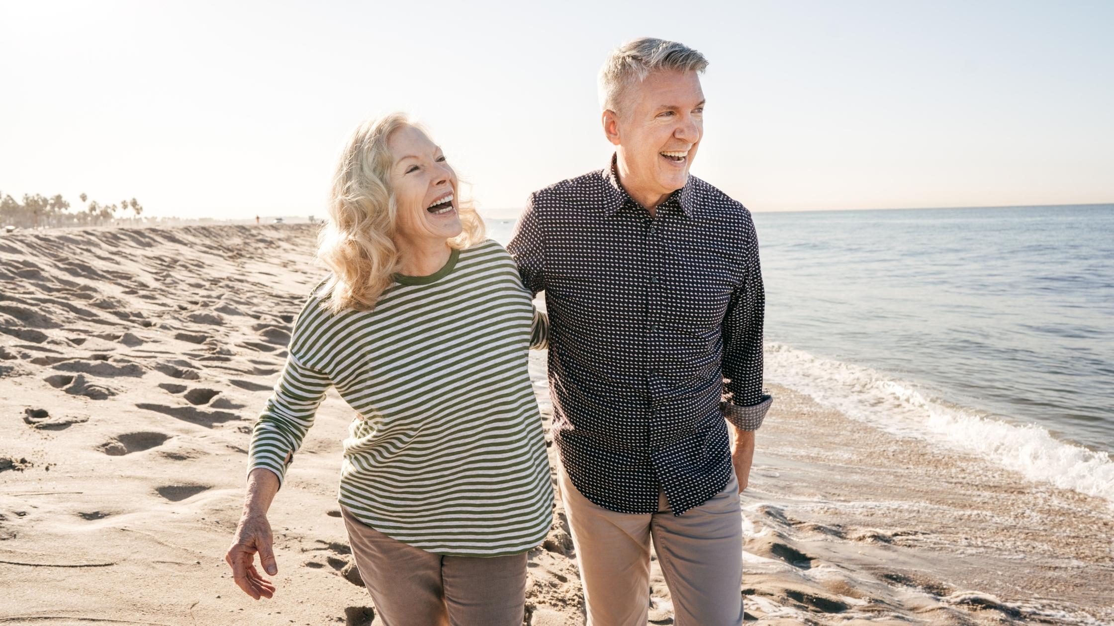 An old couple laughing and walking on the sand beside the beach.